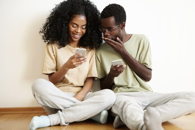 Indoor shot of stunned and shocked young dark-skinned man in glasses covering his mouth while looking at screen of his girlfriend's cell phone.