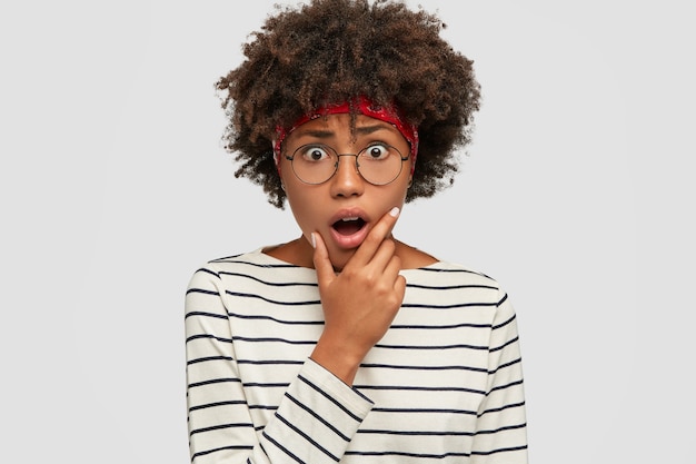 Indoor shot of stupefied black woman keeps jaw dropped, looks in terror, wears casual clothes