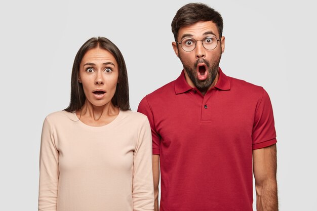 Indoor shot of stupefied young European woman and man have surprised expressions, keep mouthes opened