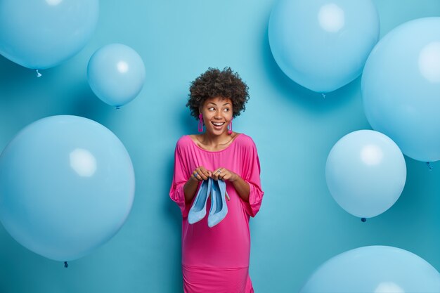 Indoor shot of stylish glad woman with Afro hair, dressed in pink dress, holds high heeled shoes, prepares for birthday party, tries to make choice what to wear, looks aside, blue balloons around