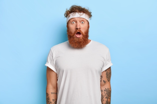 Indoor shot of surprised bearded man with red hair, stares with astonishment, has gymnastic every day