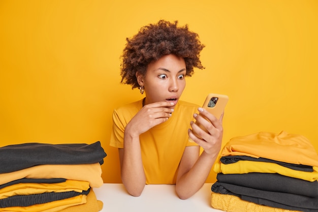 Indoor shot of surprised curly haired Afro American woman stares at smartphone display sits at table with two piles of neatly folded washed laundry isolated over yellow 