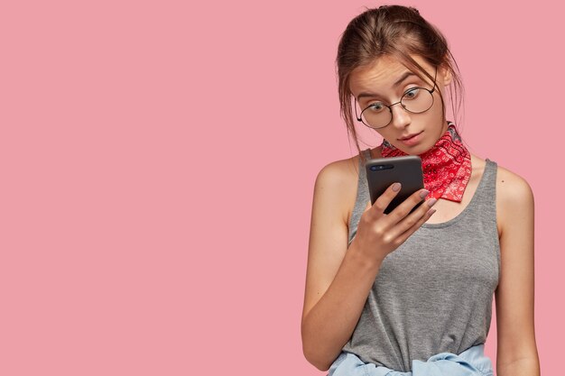 Indoor shot of surprised dark haired woman wears round transparent glasses, stares in screen of smart phone