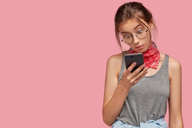 Indoor shot of surprised dark haired woman wears round transparent glasses, stares in screen of smart phone