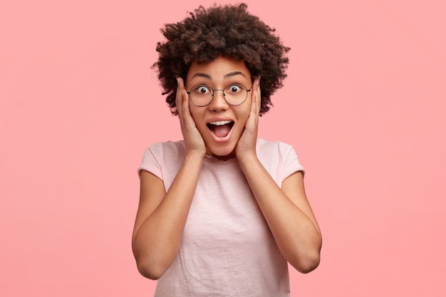 Foto gratuita ripresa in interni di una donna felice e sorpresa dall'aspetto accattivante, capelli neri ricci e pelle scura, guarda con occhi pieni di incredulità, tiene le mani sulle guance, riceve qualcosa di inaspettato e costoso