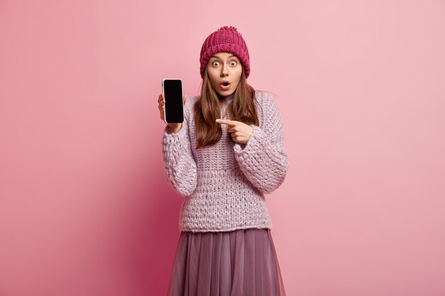 Indoor shot of surprised millenial girl shows you smartphone display, promots nice photo edit application