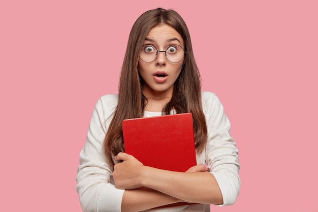 Indoor shot of surprised puzzled bugged eyed pretty schoolgirl reacts on unexpected news, holds red book