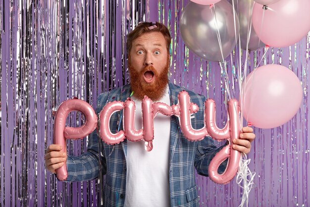 Indoor shot of surprised red haired man opens mouth from amazement, holds letter shaped balloon, wears fashionable clothes, isolated over purple wall with bright tinsel. Party time concept