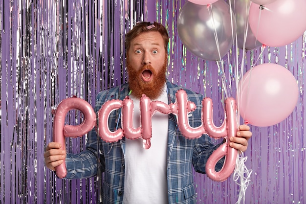 Indoor shot of surprised red haired man opens mouth from amazement, holds letter shaped balloon, wears fashionable clothes, isolated over purple wall with bright tinsel. Party time concept