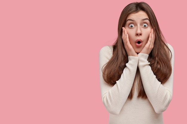 Indoor shot of surprised scared woman keeps hands on cheeks, stares with bugged eyes, hears fresh rumors or fake news, has dark hair