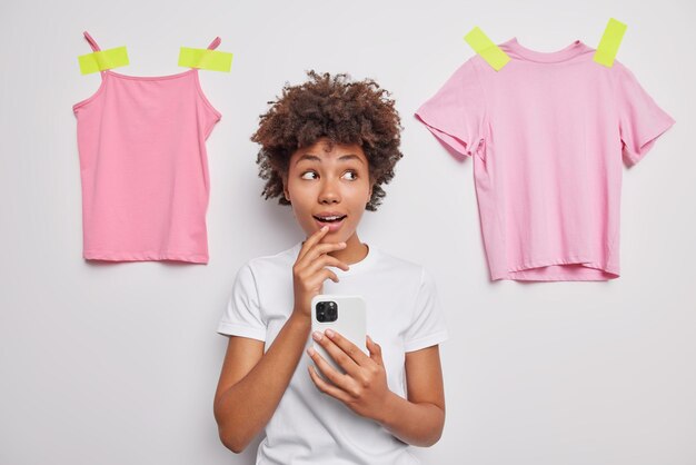 Indoor shot of surprised woman with curly hair ooks wondered aside uses mobile phones checks notifications dressed casually poses against white background with plastered pink t shirt and shirt