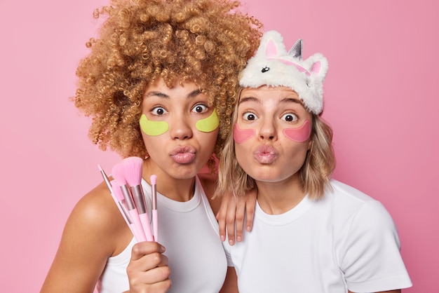Снимок в помещении: удивленные женщины держат губы сложенными, стоят близко друг к другу, держат косметические кисти, проходят косметические процедуры, наносят увлажняющие пластыри под глаза, изолированные на розовом фоне