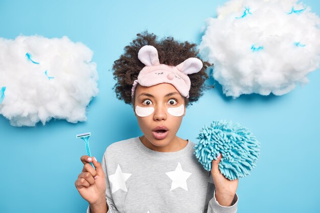 Indoor shot of surprised young Afro American woman with curly bushy hair holds razor for depilation bath sponge undergoes beauty and hygiene procedures at home after awakening in morning poses indoor