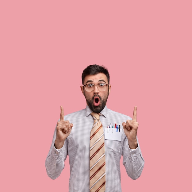 Indoor shot of surprised young entrepreneur points with both index fingers upwards, shows blank space, wears formal shirt and tie