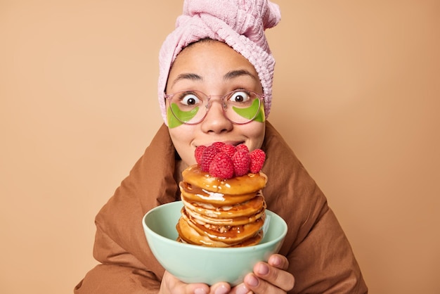 Снимок в помещении: удивленная молодая женщина смотрит в шоке, держит аппетитные блины с сиропом, носит завернутое полотенце на голове и мягкое одеяло, изолированное над бежевой стеной студии Концепция времени завтрака