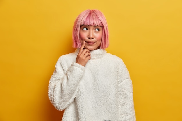 Indoor shot of suspicious thoughtful woman with eastern appearance, makes assumption, thinks about something, keeps finger near mouth, dressed in white jumper