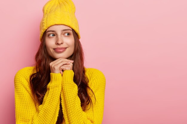 Indoor shot of tender thoughtful woman keeps hands together under chin, wears stylish yellow hat and warm sweater, poses against rosy wall, blank space.