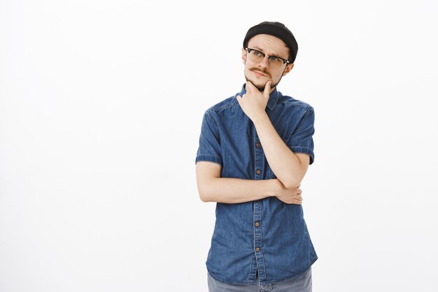 Indoor shot of thoughtful attractive adult caucasian guy in black beanie and glasses rubbing moustache on chin looking at upper left corner concentrated thinking, making decision or choice in mind