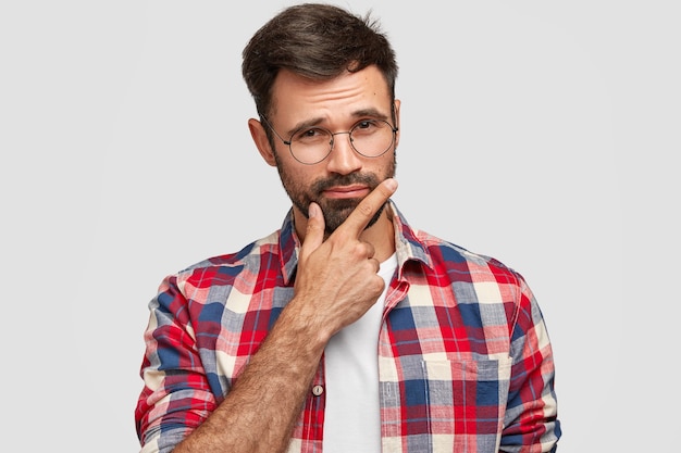 Indoor shot of thoughtful bearded young European man keeps hand under chin, looks seriously, dressed in casual checkered shirt, isolated over white wall. Masculinity concept.