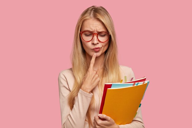 Indoor shot of thoughtful blonde college student posing against the pink wall