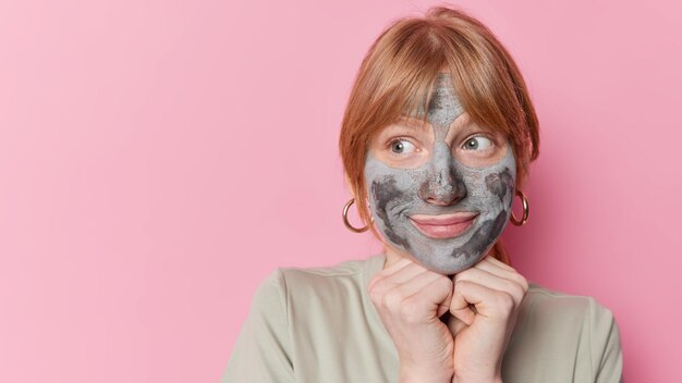 Indoor shot of thoughtful millennial girl with ginger hair applies nourishing clay mask for skin treatment keeps hands under chin looks away isolated over pink background blak space for your text