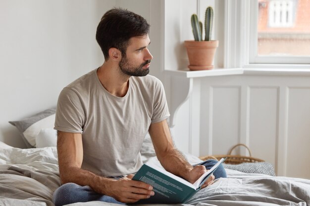 Indoor shot of thoughtful unshaven man reads books, learns some tips for successful project, sits at bed, dressed in casual clothing, focused aside, has dark stubble. Leisure and literature concept