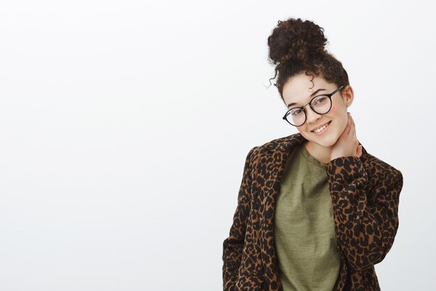 Indoor shot of timid good-looking shy girlfriend in black glasses with bun hairstyle, touching neck and tilting head with beatiful flirty smile