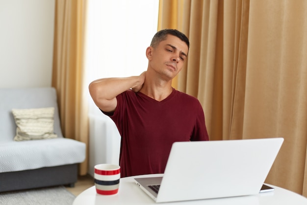 Free photo indoor shot of tired man working online at home, sitting at table in living room against sofa, having lots freelance work, having pain in neck, massaging, looking at laptop display.