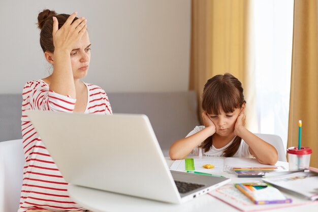 Indoor shot of tired nervous woman doing homework with daughter, keeping hand on forehead, does not know how to do task, schoolgirl sitting with palms on cheeks in front of laptop.