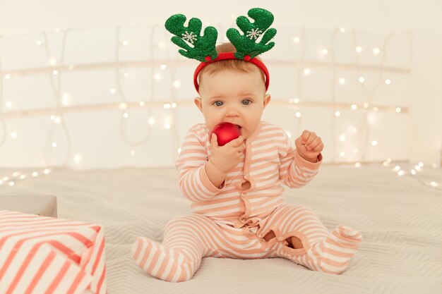 Indoor shot of toddler girl sitting on New year decorations bed and wearing striped long sleeve baby sleeper and festive deer horns, biting red Christmas ball.