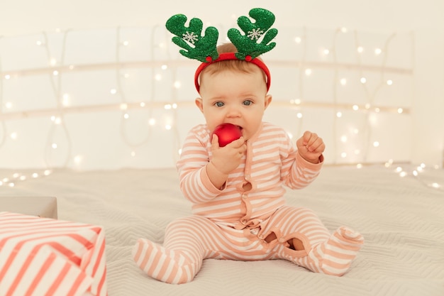 Indoor shot of toddler girl sitting on New year decorations bed and wearing striped long sleeve baby sleeper and festive deer horns, biting red Christmas ball.