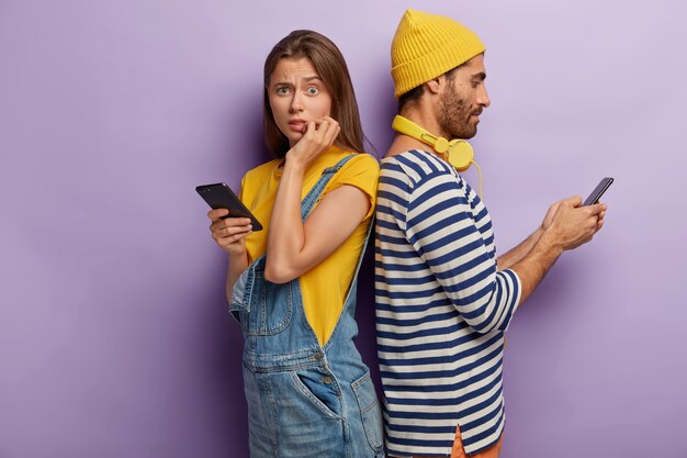 Indoor shot of two friends stand back to each other, use modern gadgets