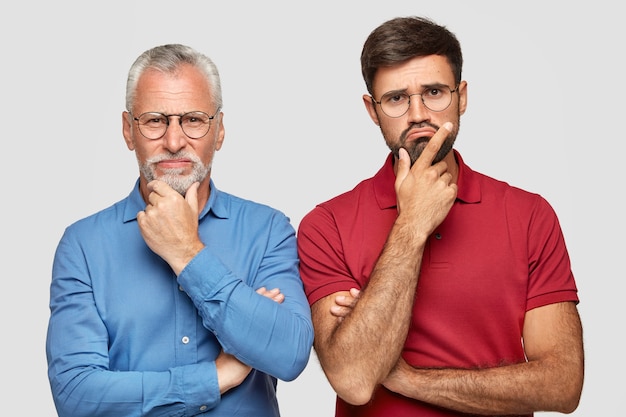 Indoor shot of two partners of different age, hold chin and look with displeased expressions, cant find solution of problem, stand next to each other, isolated over white wall. Emotions concept