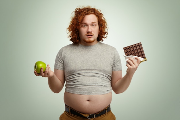 Indoor shot of uncertain confused plump young male facing hard choice as he has to chose between fresh organic apple in one hand and delicious bar of chocolate in other. dilemma, diet and food Free Photo