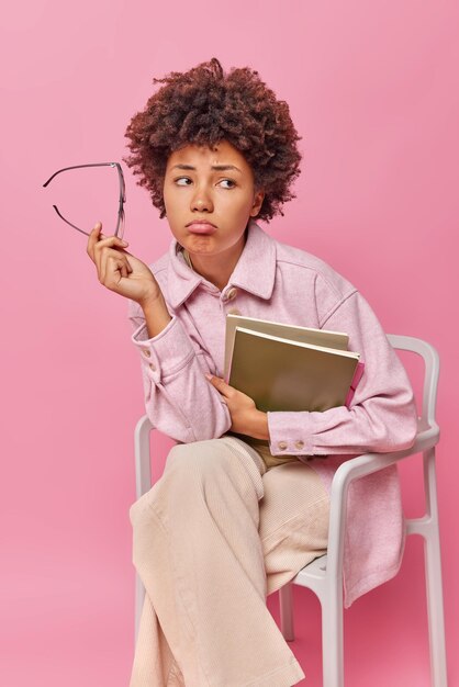 Indoor shot of unhappy female student fees tired of studying takes off glases holds textbook and notepad sits comfortably on chair wears casual clothes isolated over pink wall.