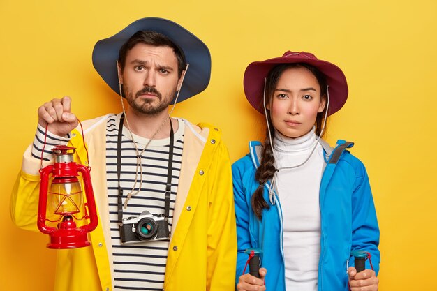 Indoor shot of unhappy mixed race woman and man stand next to each other, use kerosene lamp for lightening, trekking poles, isolated on yellow