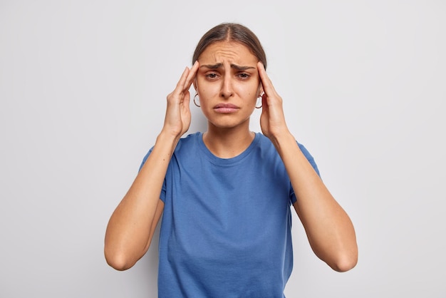 Indoor shot of unhappy tensed woman keeps hands on temples suffers from migraine stands distressed needs painkillers wears casual blue t shirt isolated over white wall
