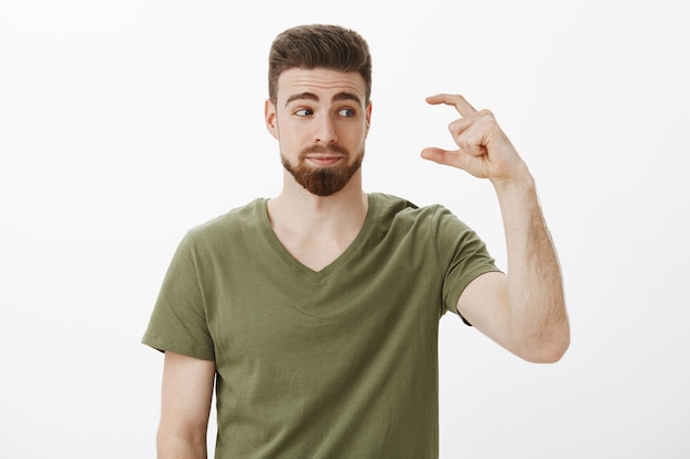 Indoor shot of unimpressed cute bearded guy waited something more shape small or tiny object looking at hand with smirk and dissatisfied look as anticipated more over white wall