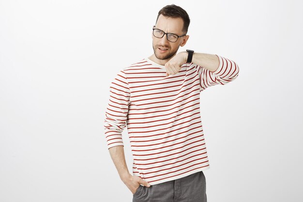 Indoor shot of unsure confused bearded guy in glasses, lifting eyebrow and grimacing while holding hand near ear and listening digital watch sound