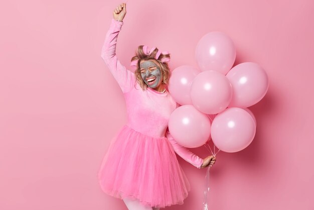 Indoor shot of upbeat glad woman dances carefree has good mood foolishes around on party holds helium balloons undergo beauty procedures wears dress isolated over pink background People and holidays