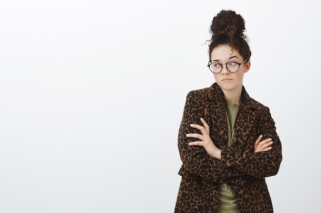 Indoor shot of upset jealous trendy girlfriend in black glasses and leopard coat over t-shirt, crossing hands on chest and looking left with bothered offended expression