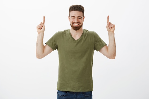 Indoor shot of very happy handsome bearded boyfriend cheering and feeling joyful smiling giggling with close eyes as pointing with raised index fingers upwards against white wall