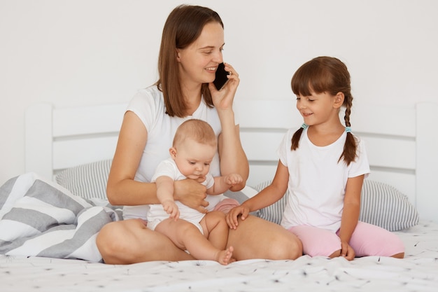 Indoor shot of winsome woman wearing white casual style t shirt sitting on bed with her children and talking via cell phone, having positive facial expression.