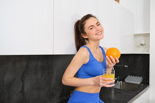 Free photo indoor shot of woman after workout standing in kitchen with fresh juice and an orange drinking it