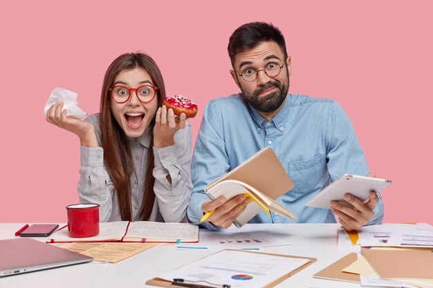 Indoor shot of woman and man coworkers eat tasty doughnut, write down notes in notebook, use modern technologies