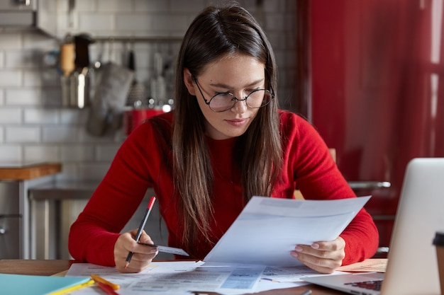 Indoor shot of woman in optical glasses checking bank account, receiving bills, holding plastic card