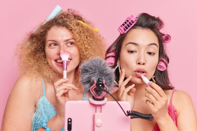 Indoor shot of women bloggers talk about makeup use cosmetic brush apply lipstick give advice to followers how to be beautiful record video via smartphone isolated over pink studio background