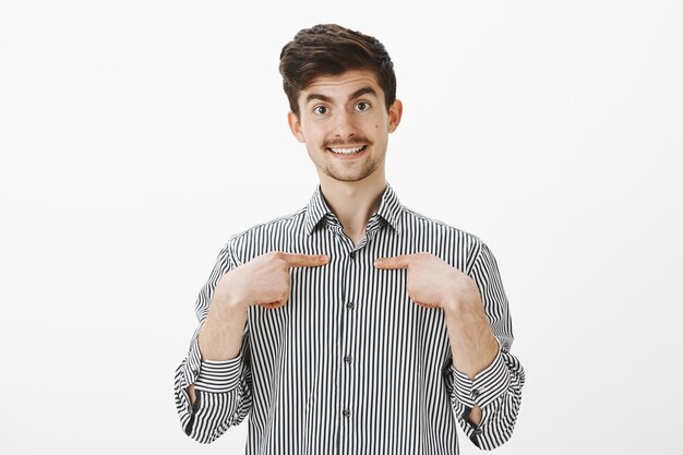 Indoor shot of wondered surprised happy friendly man in striped shirt, pointing with index fingers at chest and smiling broadly, being picked by classmates to represent group in student council