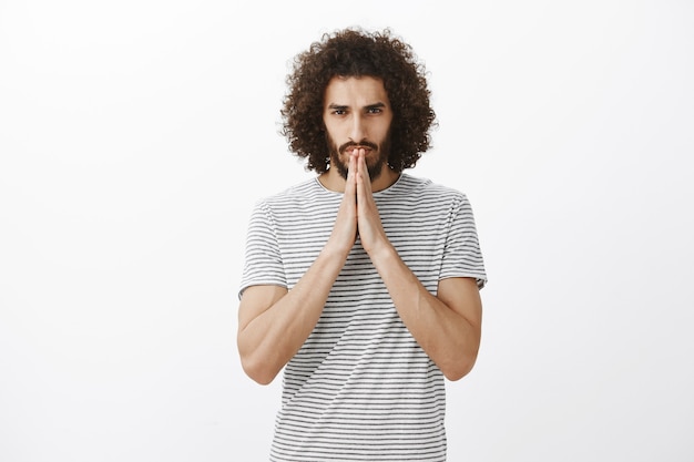 Indoor shot of worried hoping man with beard and curly hair, holding hands in pray over mouth