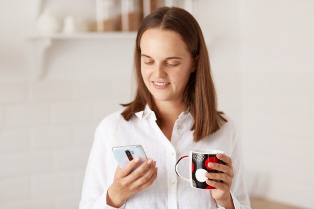 Indoor shot of young adult beautiful woman using cell phone and having a coffee in the kitchen, holding cup of beverage in hands and looking at device display.